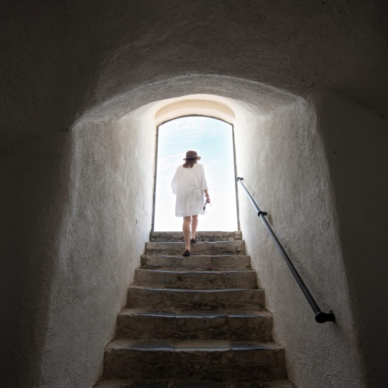 Young woman walking up a tall steep flight of stairs towards the exit and sunlight at the top in an old building viewed from the bottom of the steps